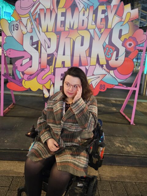 A Photodump from Wembley StadiumVD:
Image 1: A white woman with brown hair, wearing a checked jacket and sitting in a wheelchair in front of a sign that says 'Wembley sparks fly'.
Image 2: I am stood in front of a sign, that says 'so long, London' with a photo of Taylor Swift.
Image 3: A white woman, with brown wavy hair, wearing a black top and smiling at the camera. Next to her is a white man, with brown hair, wearing a black top, smiling at the camera.
