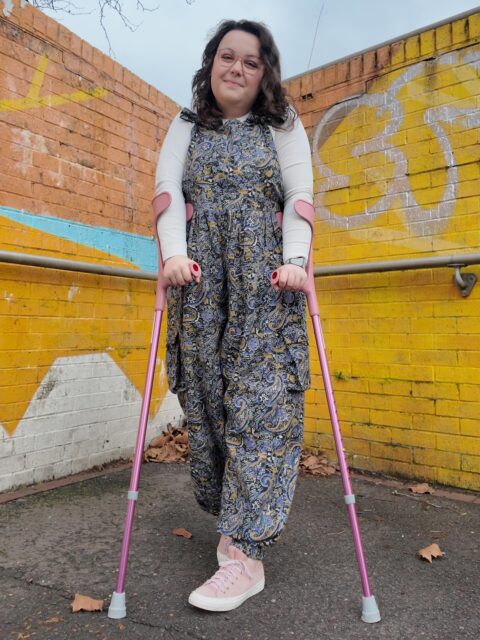 Just a Barbie girl in an inaccessible world. ✨This doll’s pink accessories are from CareCo, alongside my stunning  dungarees from Lucy and Yak!*Not sponsored, purchased with my own money.VD: A white woman, with brown, curled hair standing against a wall of abstract graffiti. She is wearing a paisley dungarees set, with pink trainers and pink crutches.