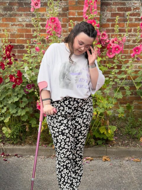 Normalising being a Swiftie who loves secondhand shopping and has matching mobility aids to her outfits 🫶🏻VD: A white woman with brown curled hair, looking downwards. She is wearing a white Taylor Swift T-shirt which says ‘Cry my eyes violet’ and black and white floral trousers. In her right hand she has a pink crutch.