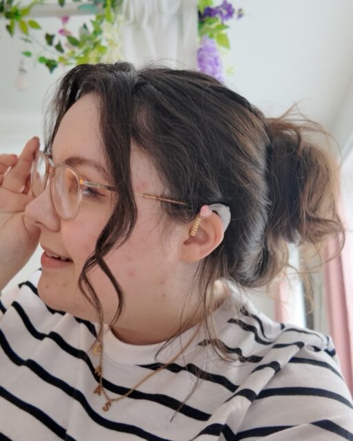 Simple hearts on my hearing aids today, I love being able to accessorise them to match my mood 🫶🏻Hearing aid accessories from @charmed.sense.accessoriesVD: A white woman with brown curly hair in a bun, wearing glasses. She is looking to one side and you can see her hearing aid with a pink to yellow ombré twist on it and a heart shaped charm.