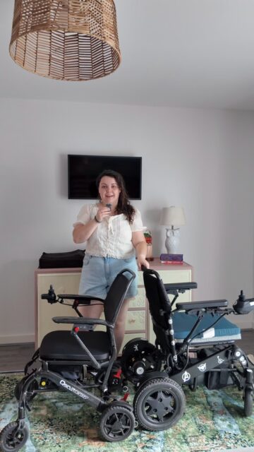 Episode 6 of The Wheel Deal: Can I lift it? Q50 wheelchairsVD: A white woman with brown hair, wearing a white top and jean shorts. She is standing next to two black folding powerchairs.