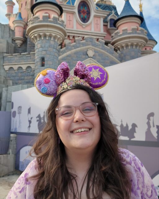 The Disney makeup of dreams... ✨ Which look is your favourite?
I truly felt like a princess every single day. 🏰VD -
Image 1: A white woman with brown wavy hair,  wearing lilac Rapunzel inspired Disney ears that sparkle. I also have a gold tiara on. I am smiling at the camera, wearing pink glasses and a lilac dress. In the background is the pink DLP castle.
Image 2: A sideways view of me, a white woman, with brown hair in a half up, half down style. I am wearing black Adventure World ears, with colourful gradients all over in sparkle. You can see my black Minnie earrings, with face glitter and hand drawn Mickey ear freckles with my pink glasses.
Image 3: A front view of the previous makeup look, now with the face glitter more obvious and my gingham red check top in view. I have taken the ears off in this selfie.
Image 4: My hair is in plaits, by my sides with orange Mickey pumpkin earrings visible. I have blue snowflake eyeshadow and a blue top.
Image 5: A close up of the silver glitter on my face and the snowflake I drew on my eyes. Pumpkin earrings in shot.