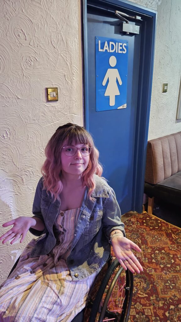 Woman in wheelchair sat outside of inaccessible ladies toilet door