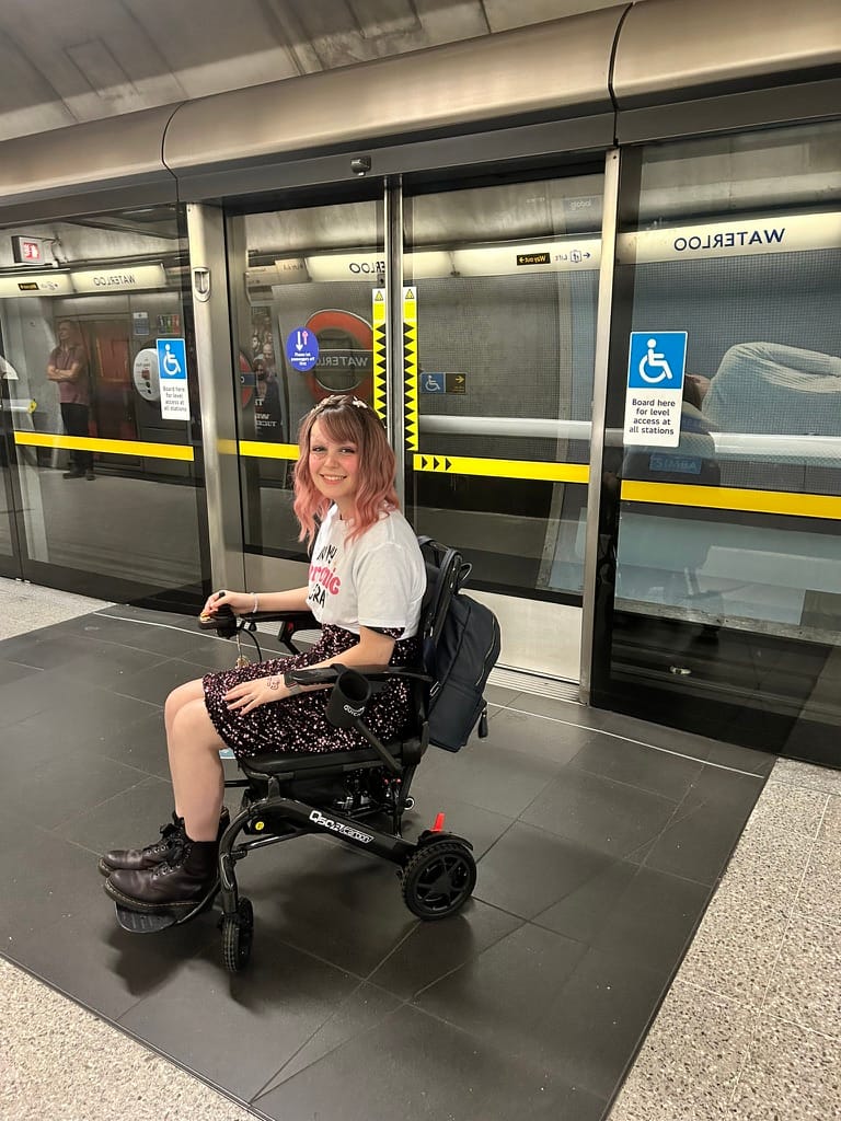 Woman with pink hair, sat in wheelchair in London underground showing off Q50r wheelchair