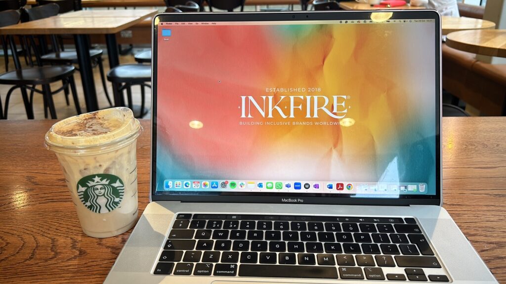 Laptop sitting on table at Starbucks with branded coffee cup sat next to it. Wallpaper has Inkfire logo against a multu-coloured background.