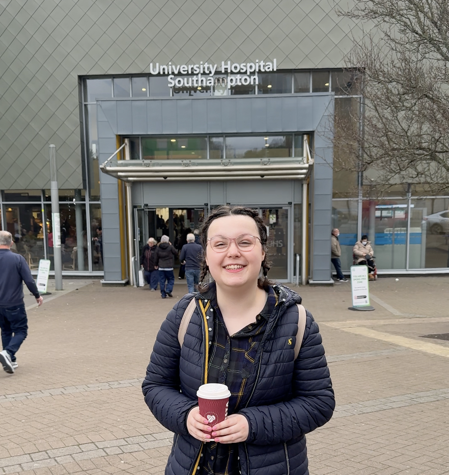 A young woman with brown hair in plaits, wearing a blue coat, holding a cup of coffee. She is stood outside of Southampton General Hospital where she receives care.